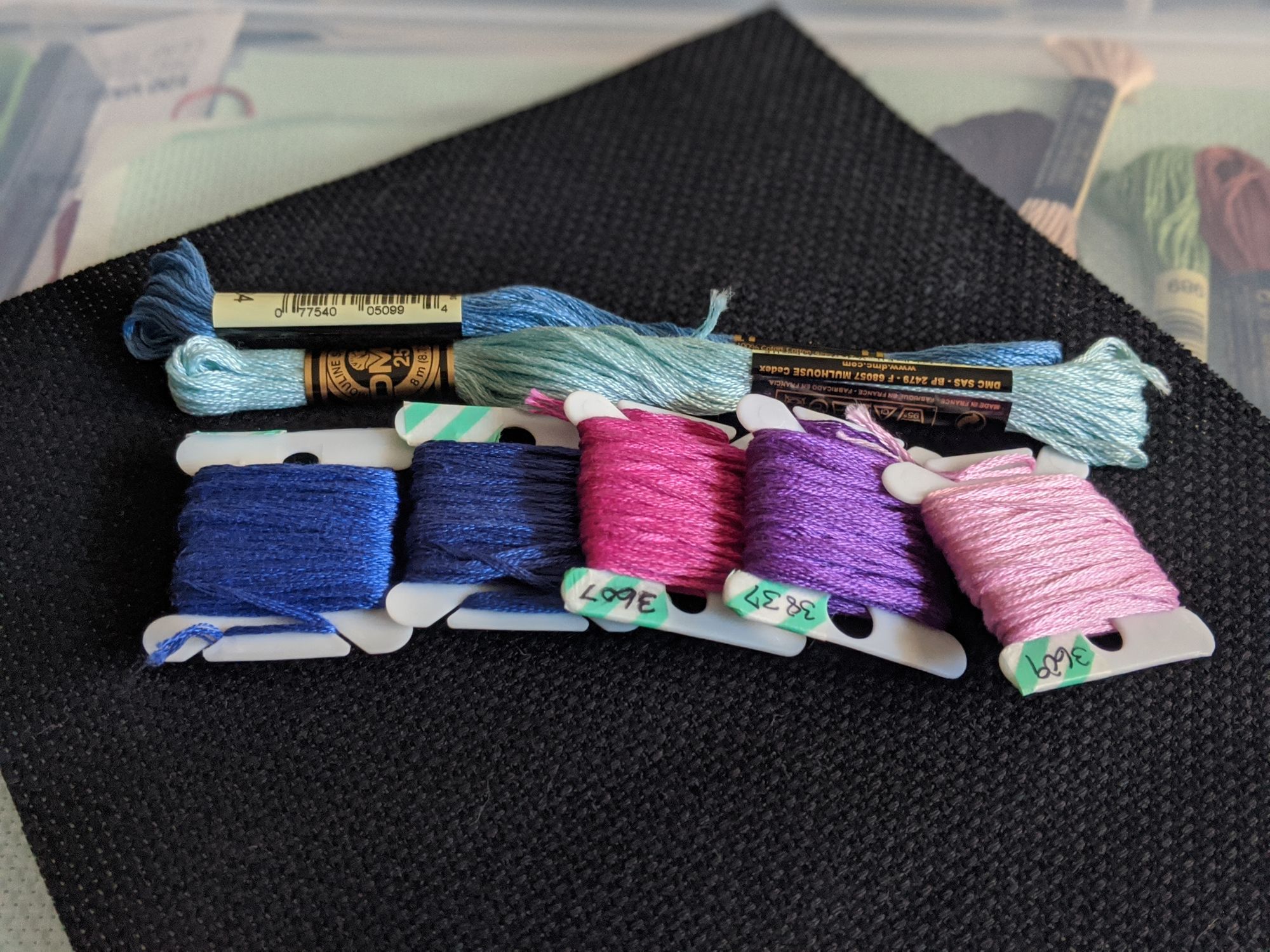 Different colored blue, pink, and purple thread sitting on top of black aida cloth, which is fabric you use for cross stitching