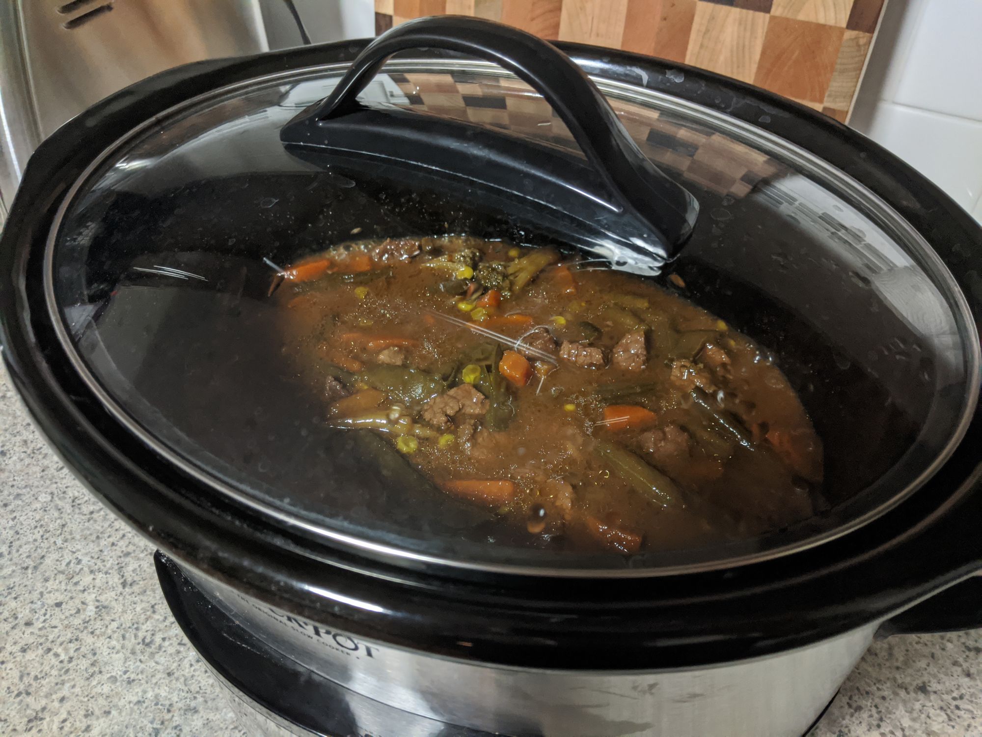 A picture taken of a slow cooker from the top view. You can see through the glass cover inside, where a beef stew looking meal is cooking away.