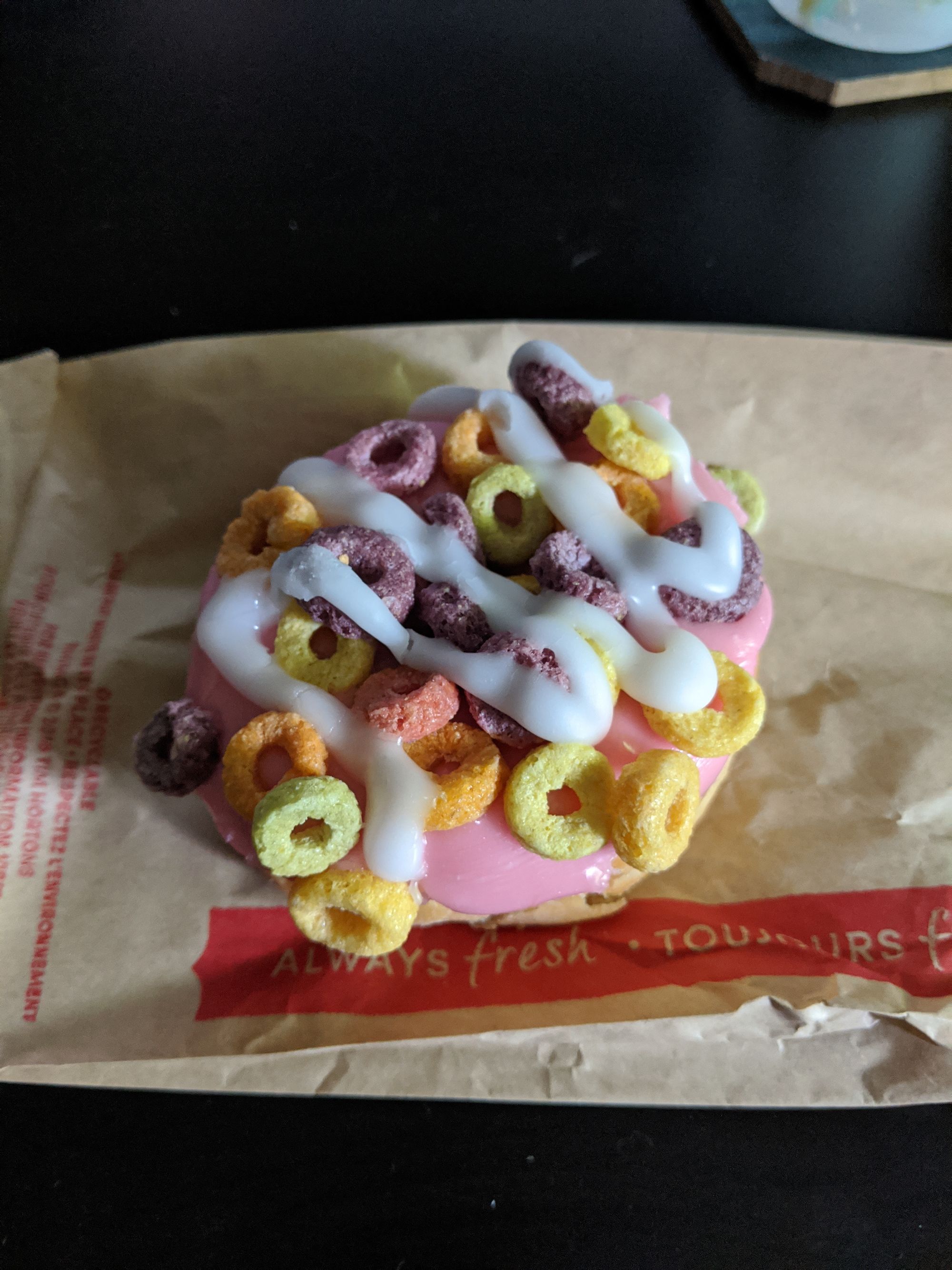 A donut with pink icing, topped with fruit loop piece, and a final drizzle of white icing on top.