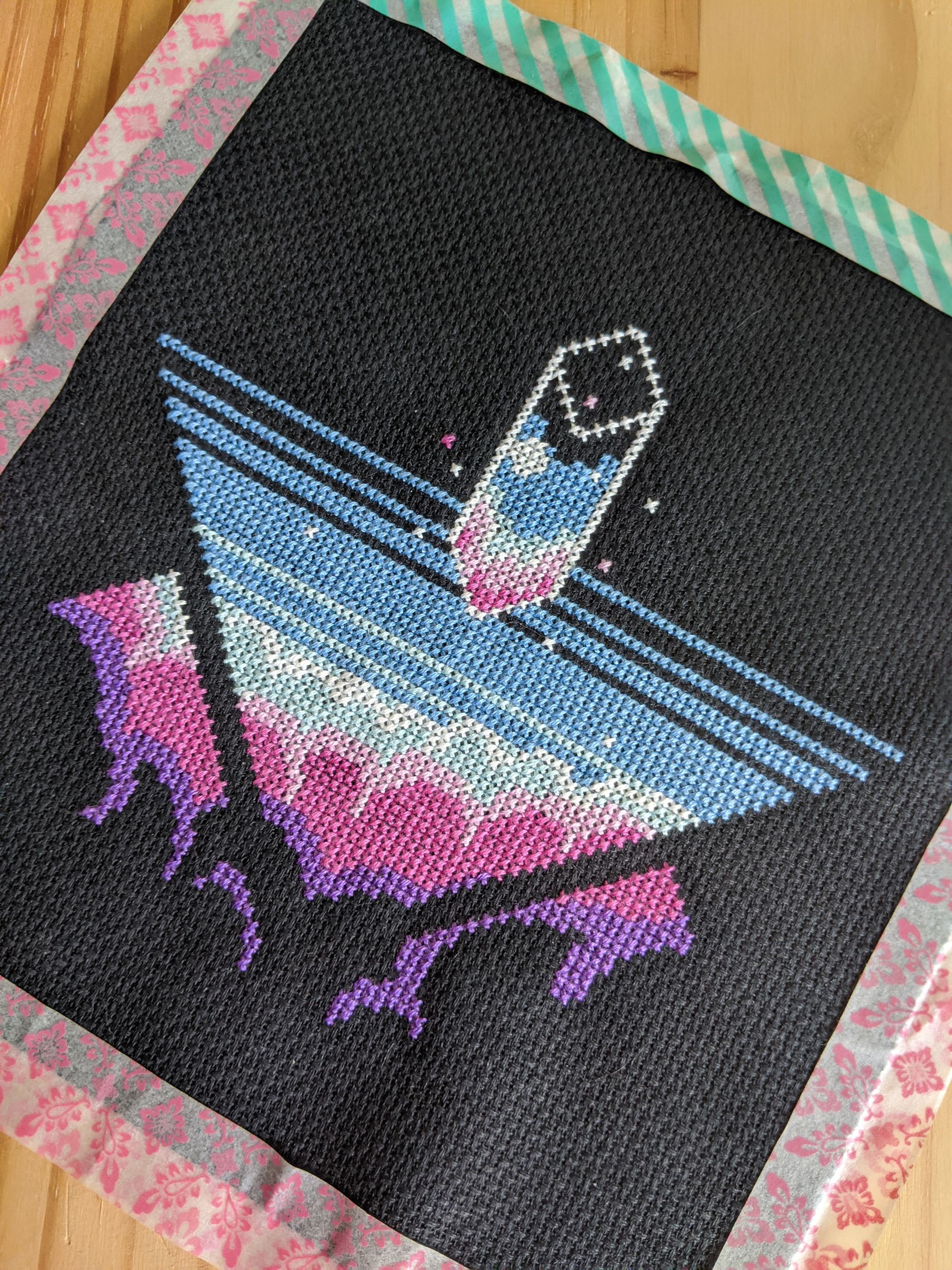 A cross stitch piece with a variety of blues, pinks and purples. It's an abstract piece with the beginnings of what looks like clouds at the bottom.