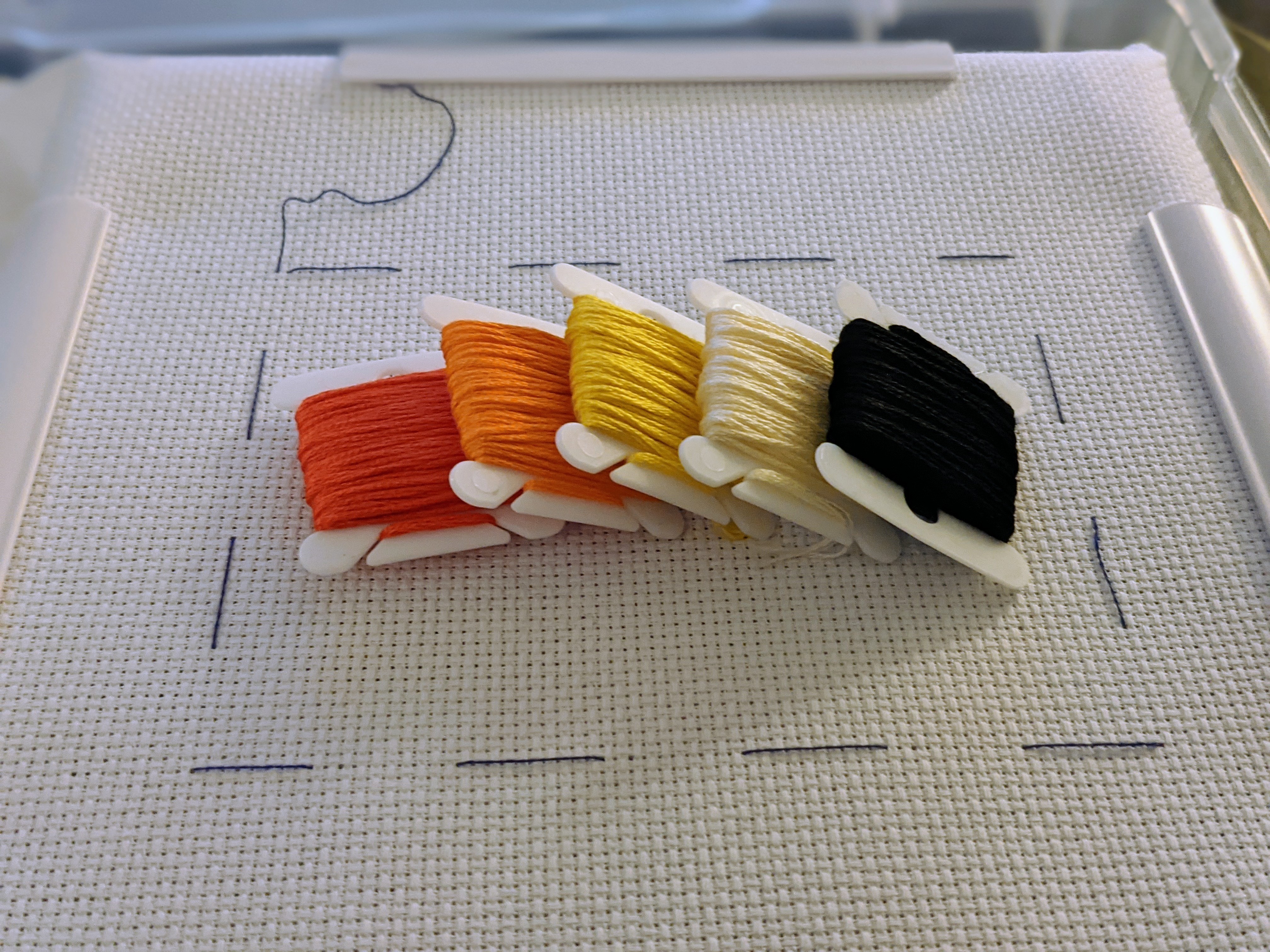 White aida cloth in a plastic Q Snap Frame, 4 colored bobbins sitting on top of it: red, orange, yellow, creme, and black.