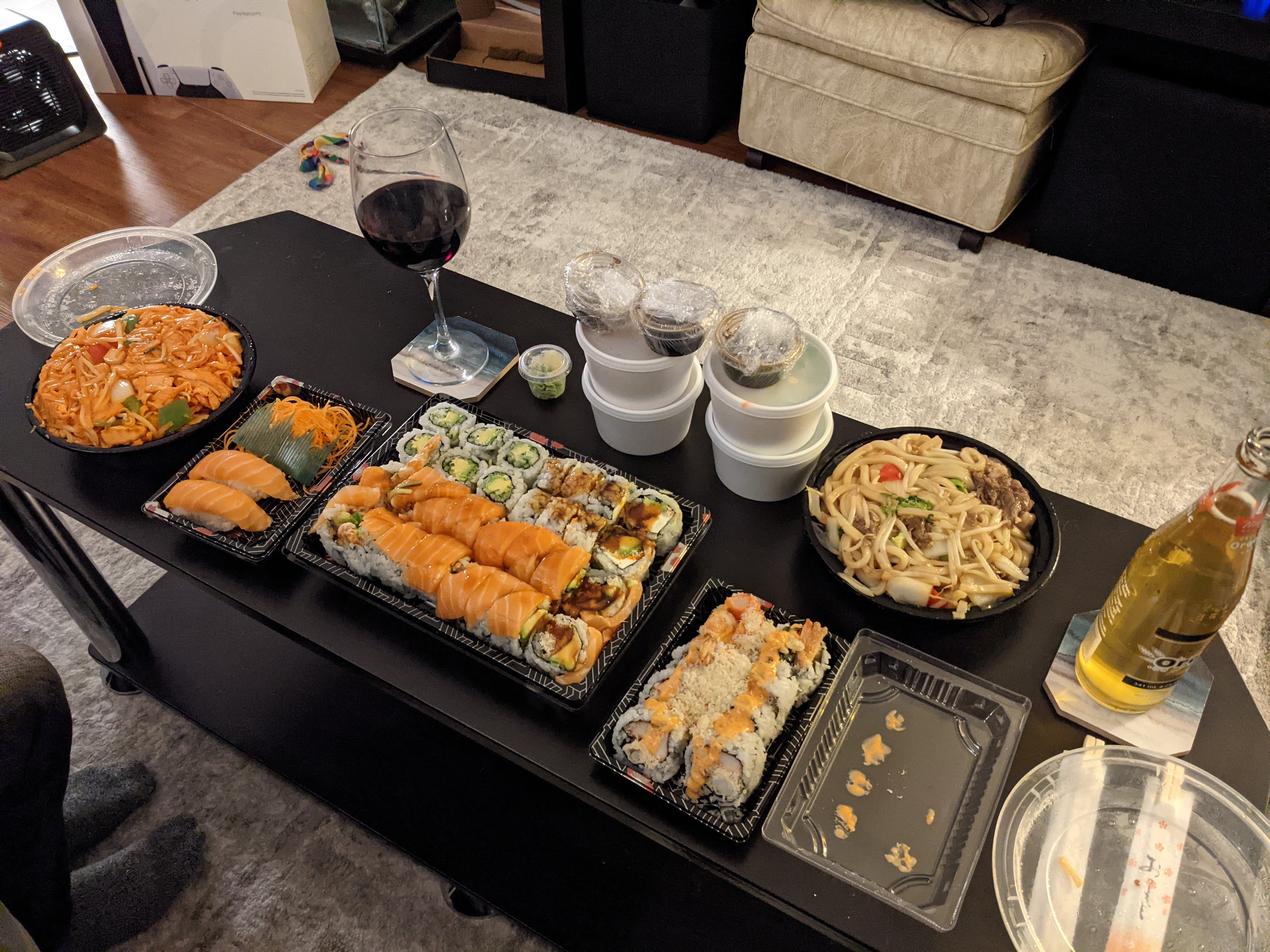 A variety of sushi dishes layed out on a black coffee table. It's lots of food with a beer and wine glass as well.