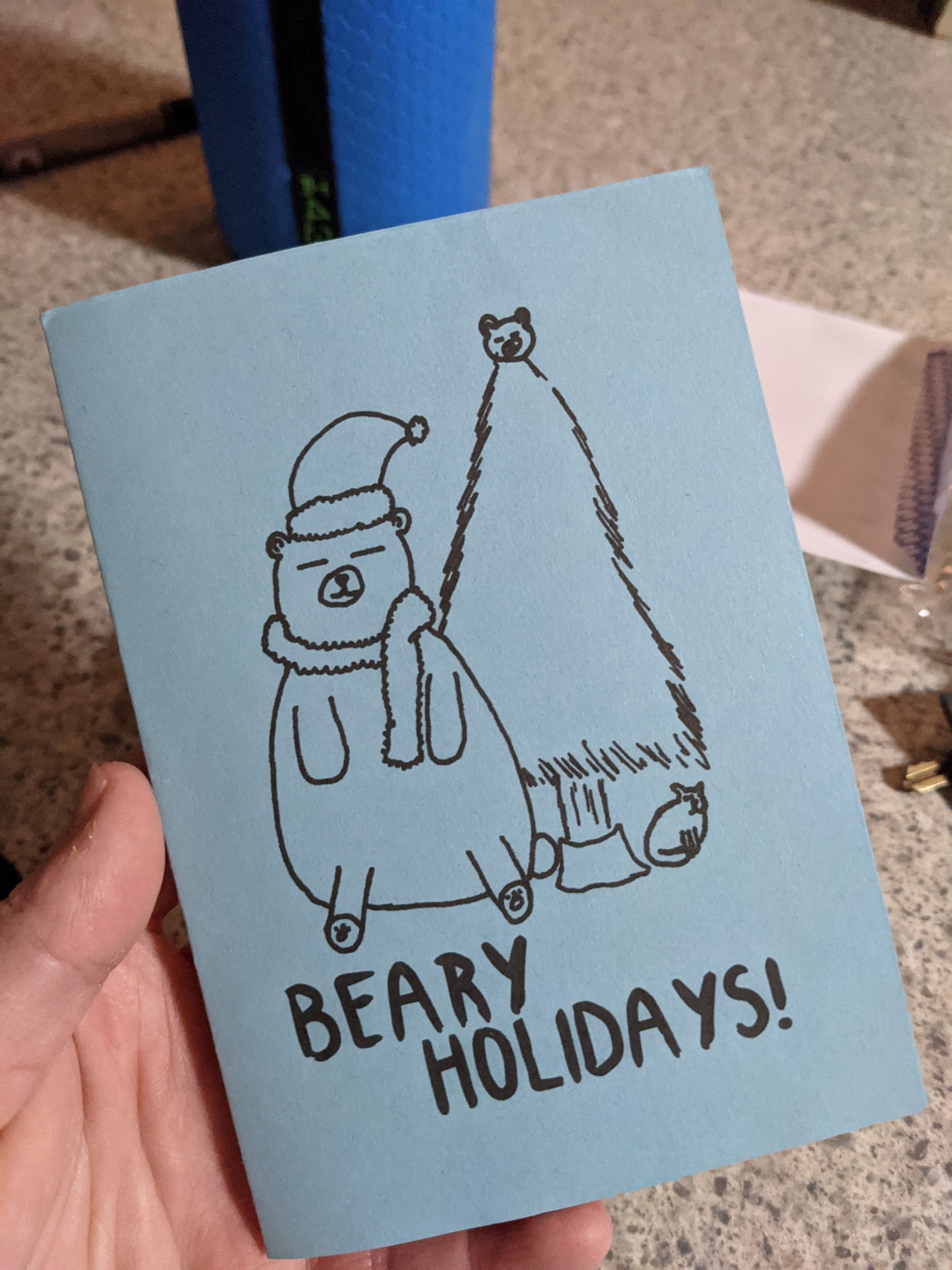 My hand holding a blue christmas card, is says Beary Holidays! with a drawn bear next to a christmas tree.