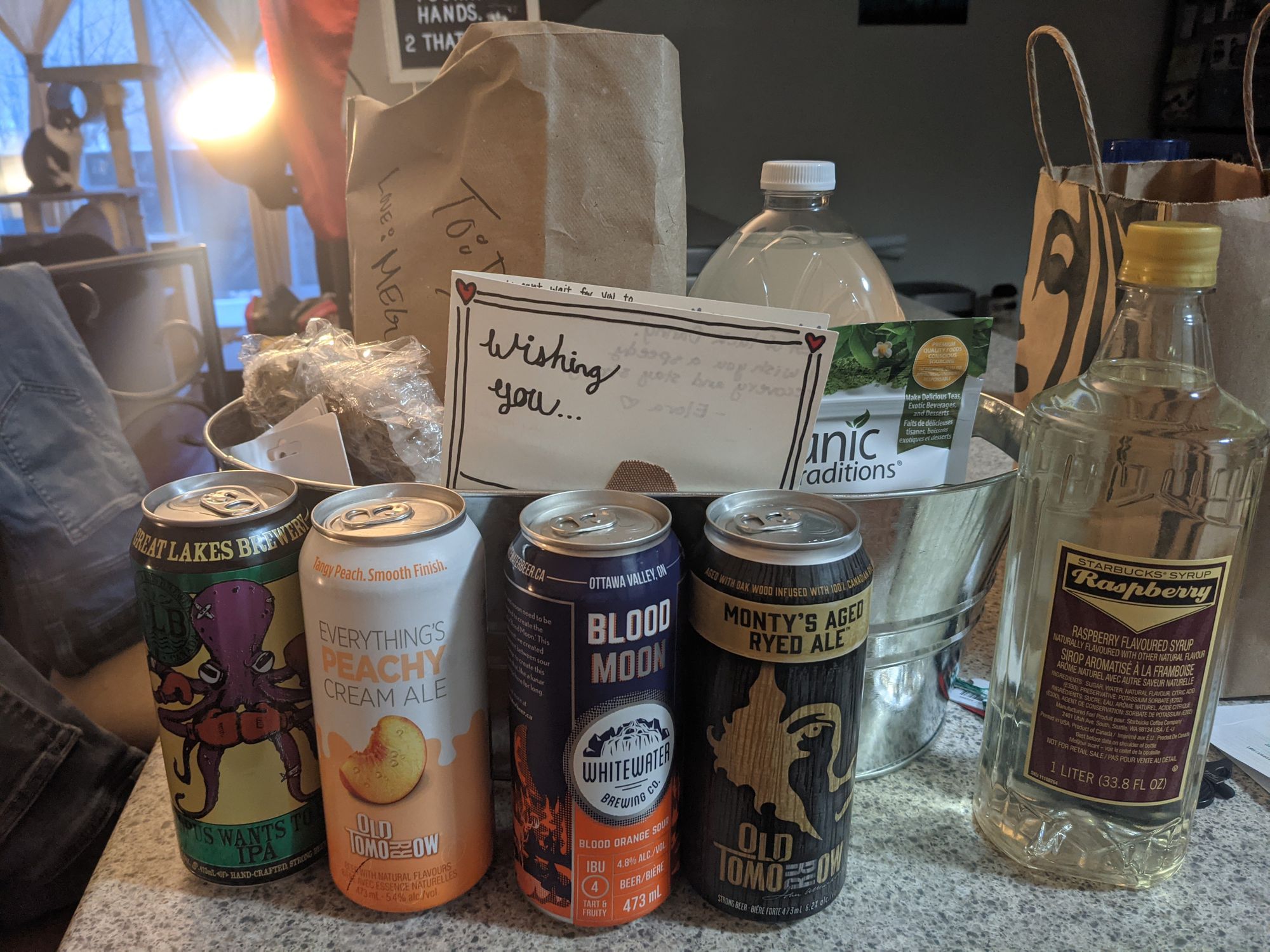 A gift basket with various items: four individual cans of beer & cider, raspberry sugar syrup, lemonade, matcha powder, various baked goods.